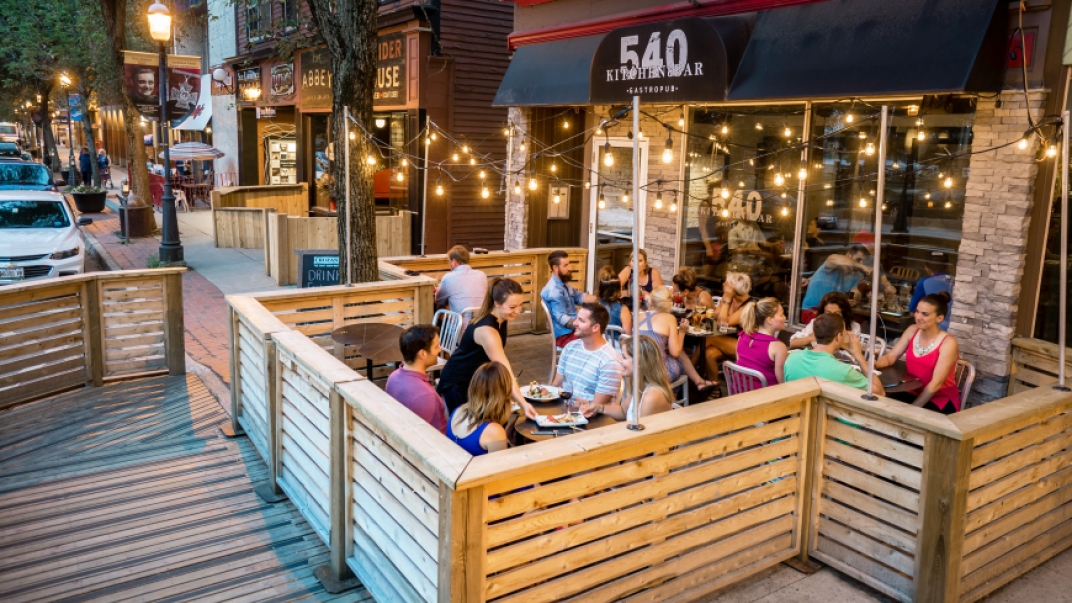 A wood patio built on a side walk. The has 4 full tables and the black awning above it reals 540 Kitchen & Bar