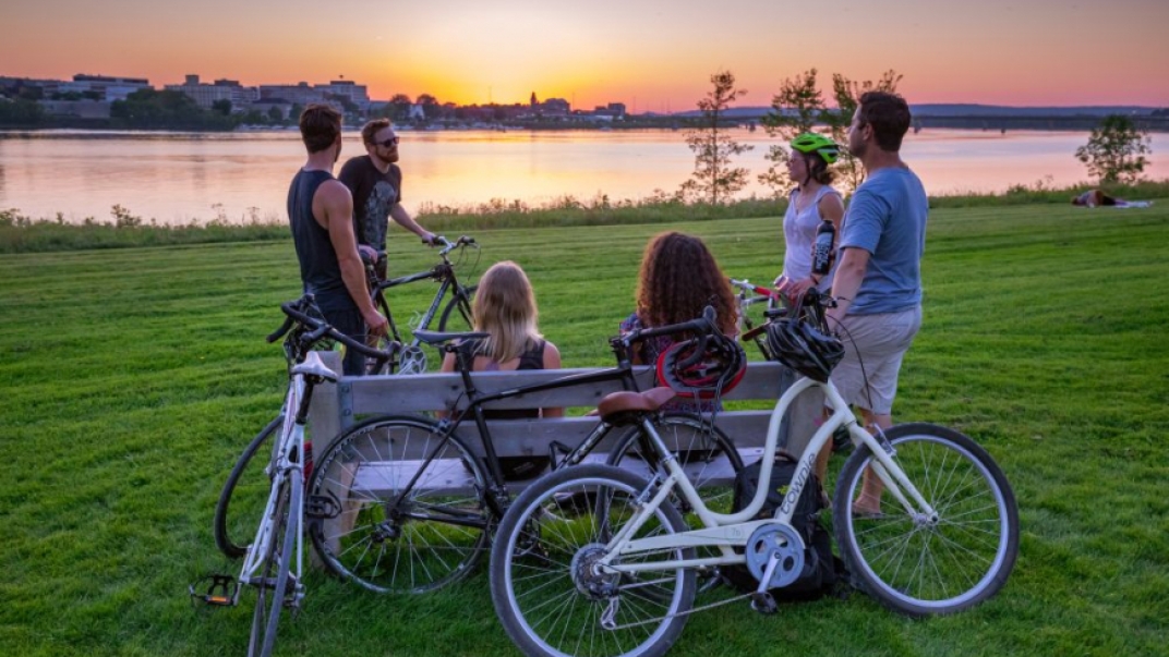 Group of people with bikes sitting on a park bench overlooking the water and city skyline