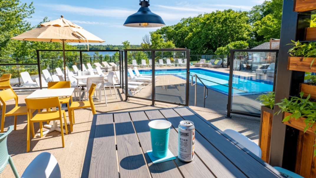 A sunny pool side. The pool is surrounded by white lounge chairs. Up a level is the dining area, with a table with a drink can on it.