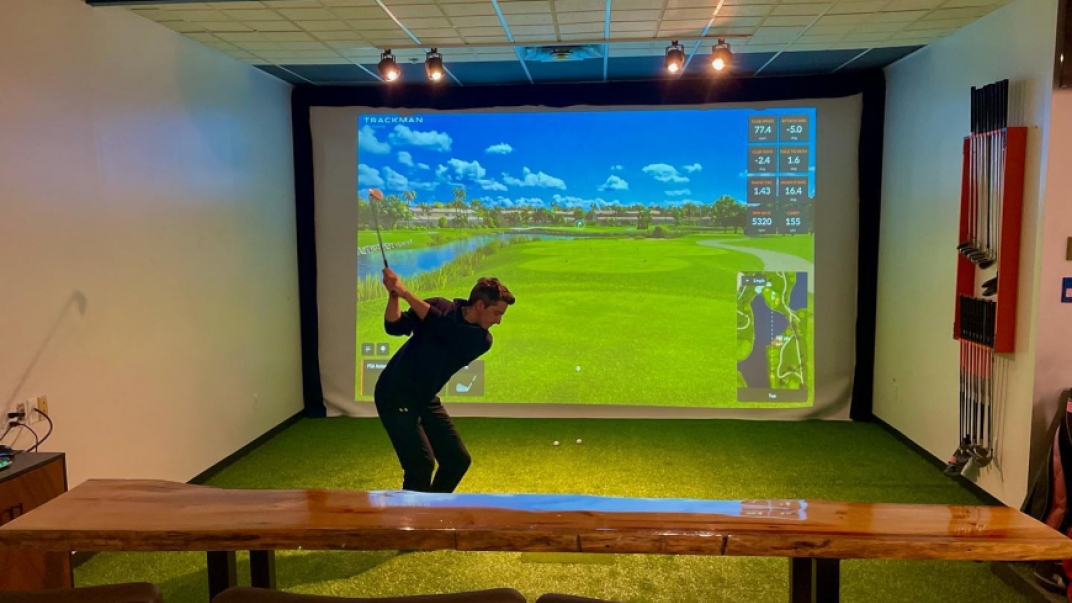 Man swinging golf club at golf ball. There is an electronic golfing projection on a screen in the background
