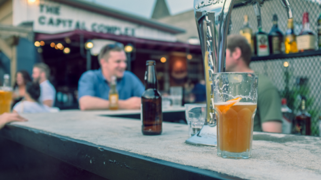 Bartop with beer bottle and pint on them. There are people conversing in the background.