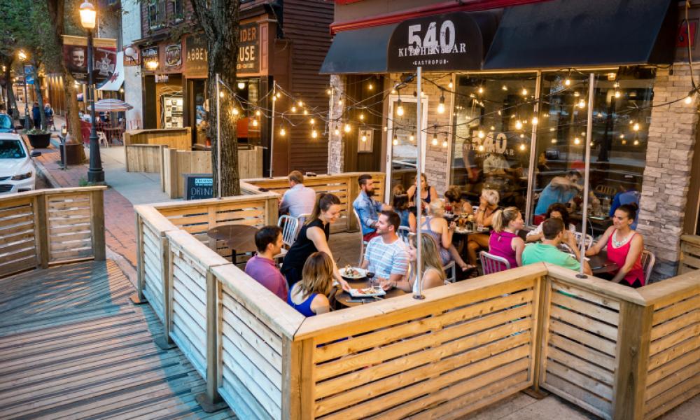 A wood patio built on a side walk. The has 4 full tables and the black awning above it reals 540 Kitchen & Bar
