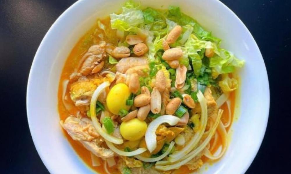 A white bowl with a yellow broth dish, featuring leafy greens, nuts, and onions.