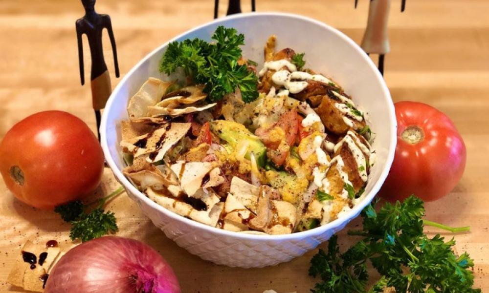 A bowl of food with pita chips crumbled into the bowl, cilantro, and meat from a kebab drizzled with sauce. There are whole tomatoes and an onion around the bowl.