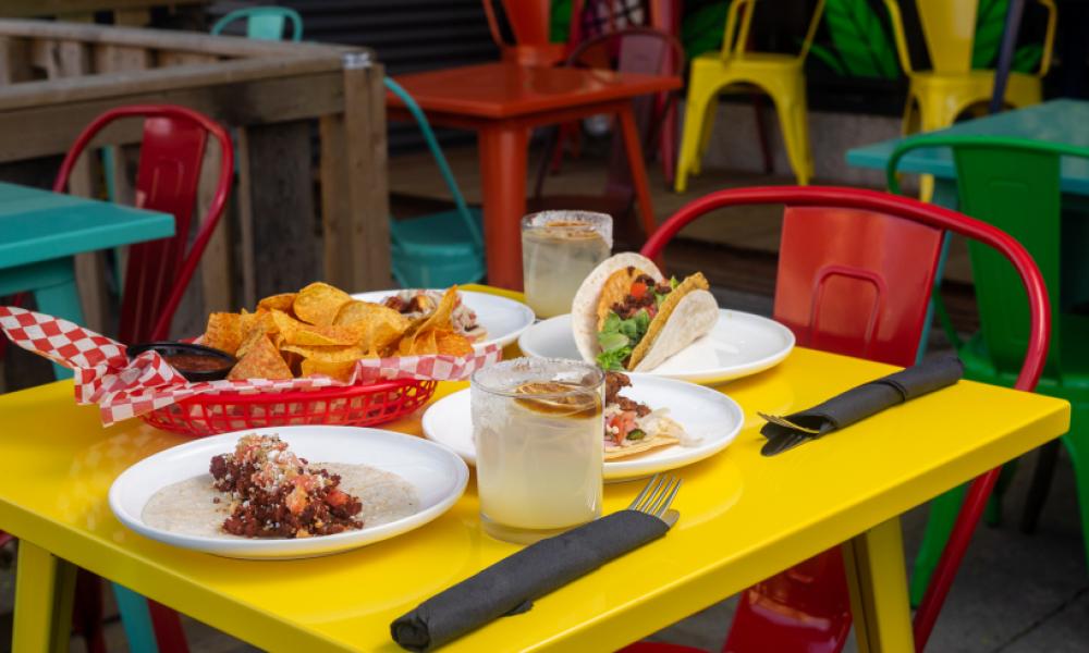 A yellow table with a red chair. There are tacos on plates on the table and a bowl of chips.