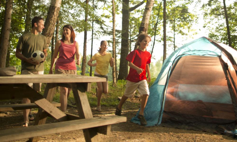 Smiling family playing at a campsite in a forest with a tent and picnic table.