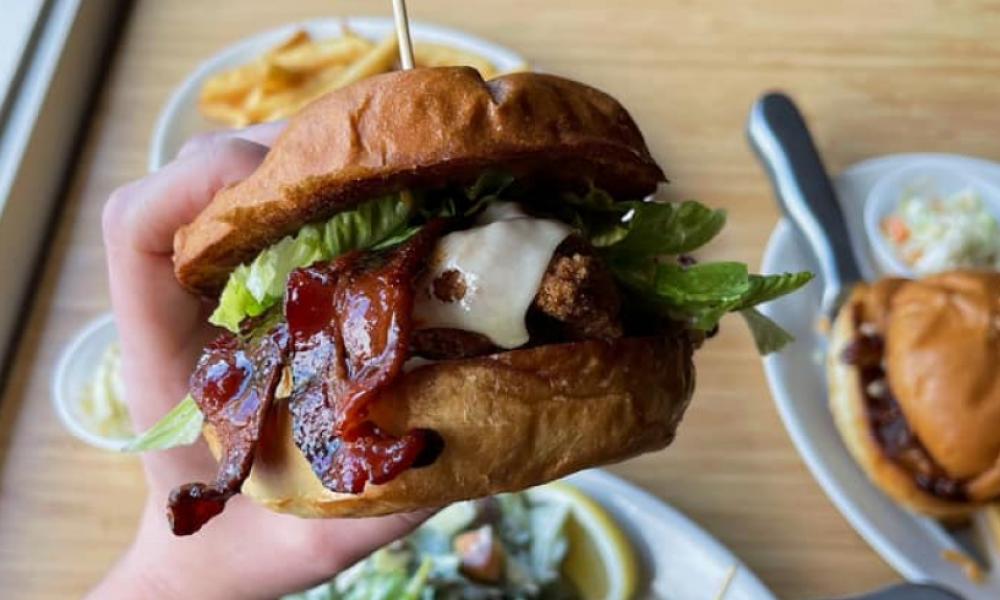 Someone holding a bacon cheeseburger close to a camera with burgers and fries on the table in the background