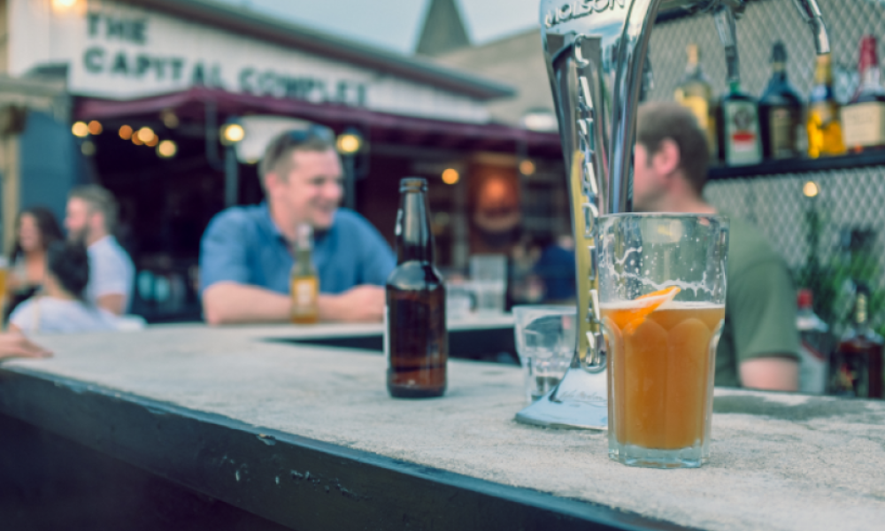 Bartop with beer bottle and pint on them. There are people conversing in the background.
