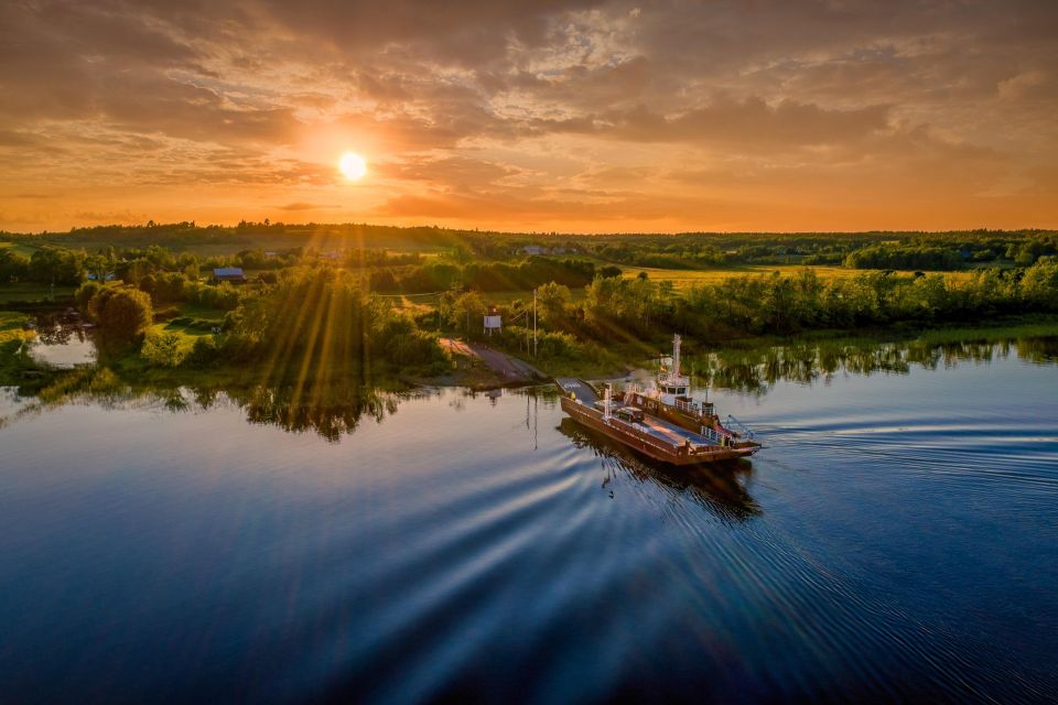 A small ferry crosses a calm river at sunset, viewed from above, with golden sunlight streaming over a green rural shoreline and the boat’s wake rippling across the water.